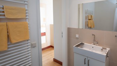Einzelzimmer mit eigenem Badezimmer mit einem weißen Waschbecken, einem Spiegel und einem Handtuchhalter mit gelben Handtüchern. Der Boden ist gefliest, und eine Tür führt zu einem angrenzenden Raum.