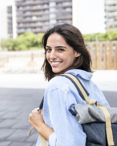 Junge Frau mit einem Rucksack, die lächelnd über die Schulter schaut. Sie trägt ein hellblaues Hemd und steht auf einem Gehweg in einer urbanen Umgebung mit modernen Gebäuden im Hintergrund.