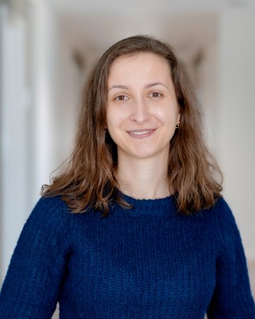 Eine lächelnde Frau mit langen, lockigen Haaren trägt einen blauen Pullover und steht in einem hellen Flur.