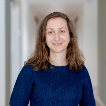 Eine lächelnde Frau mit langen, lockigen Haaren trägt einen blauen Pullover und steht in einem hellen Flur.