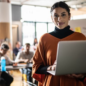 Eine Frau in einem orangefarbenen Oberteil hält einen Laptop in einem Besprechungsraum. Im Hintergrund sind mehrere Personen zu sehen, die an einem Tisch sitzen und auf sie schauen. Notizen sind an einer Wand sichtbar.