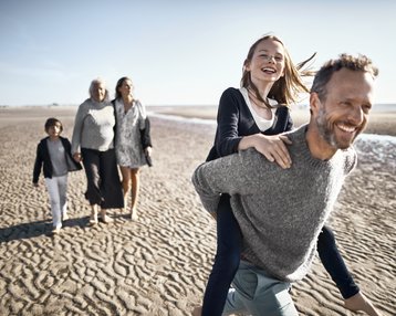 Eine Gruppe von fünf Personen geht am Strand entlang. Ein Mann trägt ein Mädchen auf dem Rücken, während zwei Frauen und ein Junge in der Nähe spazieren. Der Himmel ist klar und die Sandfläche ist sichtbar.