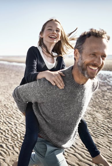 Eine Gruppe von fünf Personen geht am Strand entlang. Ein Mann trägt ein Mädchen auf dem Rücken, während zwei Frauen und ein Junge in der Nähe spazieren. Der Himmel ist klar und die Sandfläche ist sichtbar.