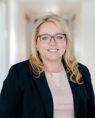 Eine blonde Frau mit Brille trägt einen schwarzen Blazer und steht in einem hellen Flur. Sie lächelt freundlich in die Kamera.