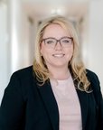 Eine blonde Frau mit Brille trägt einen schwarzen Blazer und steht in einem hellen Flur. Sie lächelt freundlich in die Kamera.