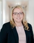 Eine blonde Frau mit Brille trägt einen schwarzen Blazer und steht in einem hellen Flur. Sie lächelt freundlich in die Kamera.