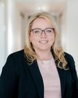 Eine blonde Frau mit Brille trägt einen schwarzen Blazer und steht in einem hellen Flur. Sie lächelt freundlich in die Kamera.