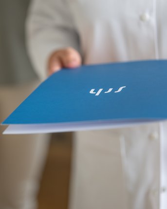 Eine Person in einem weißen Kittel hält eine blaue Mappe mit der Aufschrift "SIT" in der Hand. Im Hintergrund ist eine weitere Person in heller Kleidung zu sehen.
