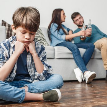 Ein Junge sitzt mit gesenktem Kopf und verschränkten Armen auf dem Boden, während ein Paar auf einem Sofa im Hintergrund miteinander spricht und Getränke in der Hand hält. Der Junge wirkt nachdenklich und abseits.