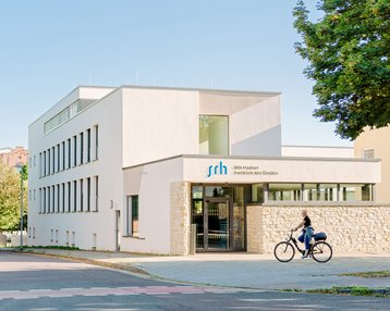 Moderne Rehaklinik für Abhängigkeitserkrankungen mit klaren Linien und hellen Farben. Eine Person fährt mit dem Fahrrad vorbei. Der Eingang ist gut sichtbar, mit dem Schriftzug "srh" über der Tür.